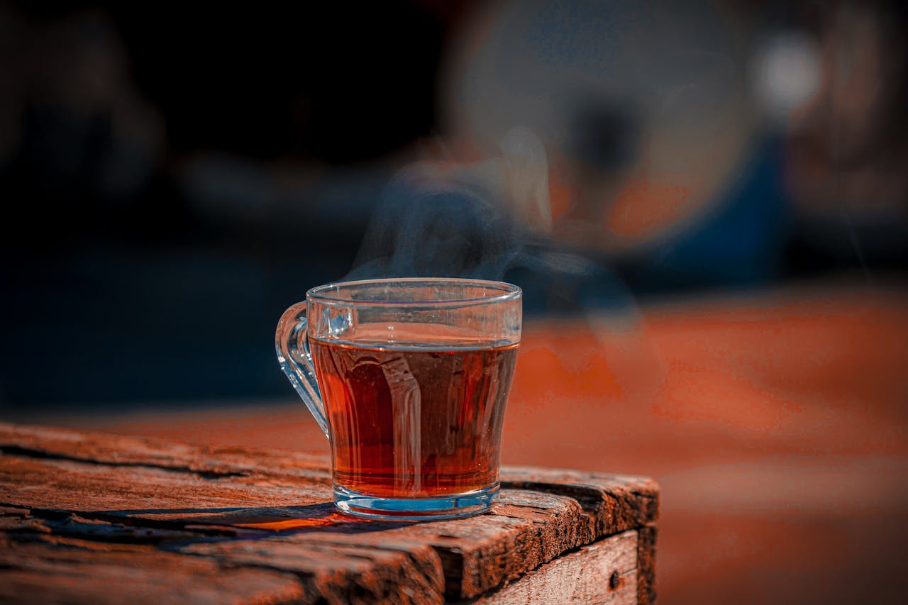 A steaming glass of tea sits on a rustic wooden table outdoors.