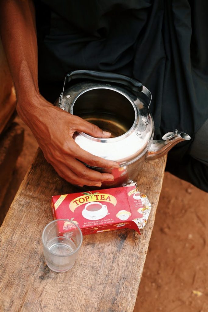 A person preparing Top Tea with a stainless steel teapot on a wooden surface, with a teabag packet and a glass.