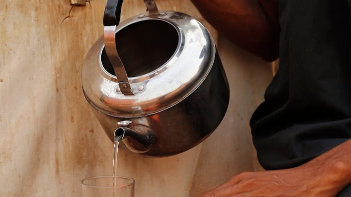 A person pours tea from a kettle into a glass outdoors in Abuja, Nigeria, with biscuits nearby.